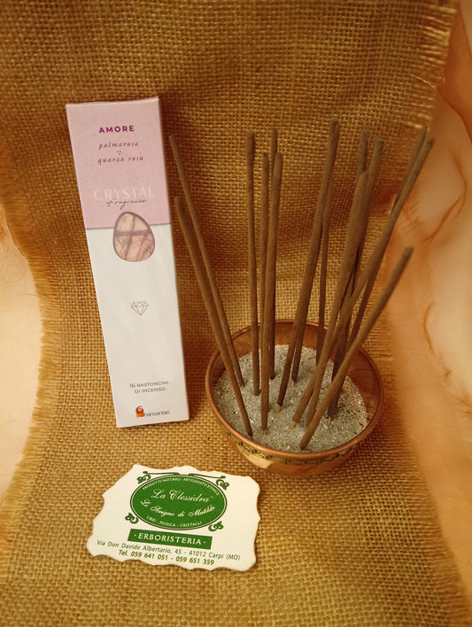 Confezione di Incenso Crystal Fragrance Amore Dhanvantari appoggiata accanto a una ciotola di rame con sedici bastoncini inseriti nella sabbia e al biglietto da visita dell’Erboristeria La Clessidra Carpi su sfondo in telo di yuta