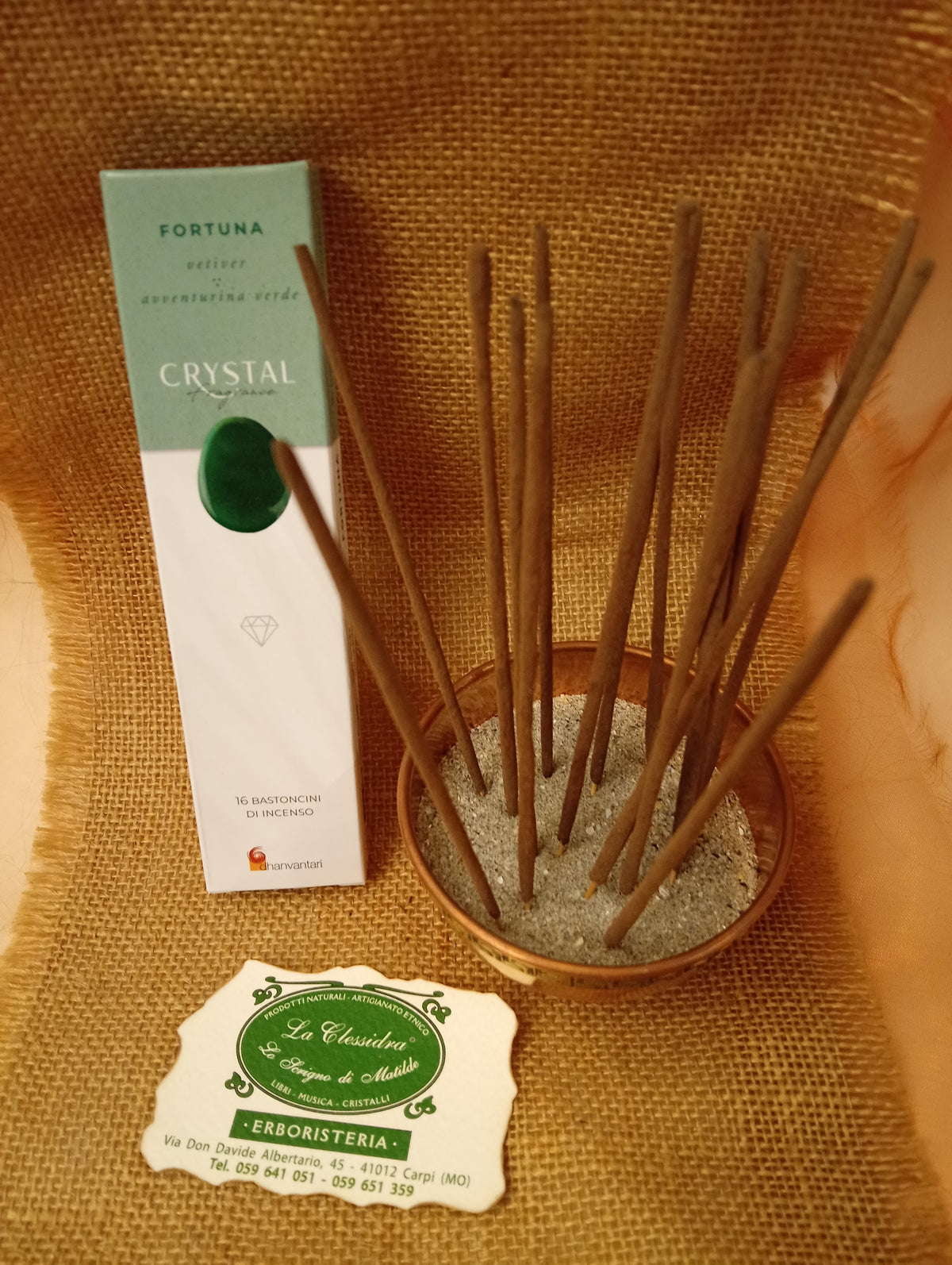 Confezione di Incenso Crystal Fragrance Fortuna Dhanvantari appoggiata accanto a una ciotola di rame con sedici bastoncini inseriti nella sabbia e al biglietto da visita dell’Erboristeria La Clessidra Carpi su sfondo in telo di yuta.