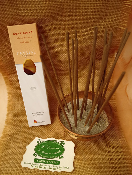 Confezione di Incenso Crystal Fragrance Guarigione Dhanvantari appoggiata accanto a una ciotola di rame con sedici bastoncini inseriti nella sabbia e al biglietto da visita dell’Erboristeria La Clessidra Carpi su sfondo in telo di yuta