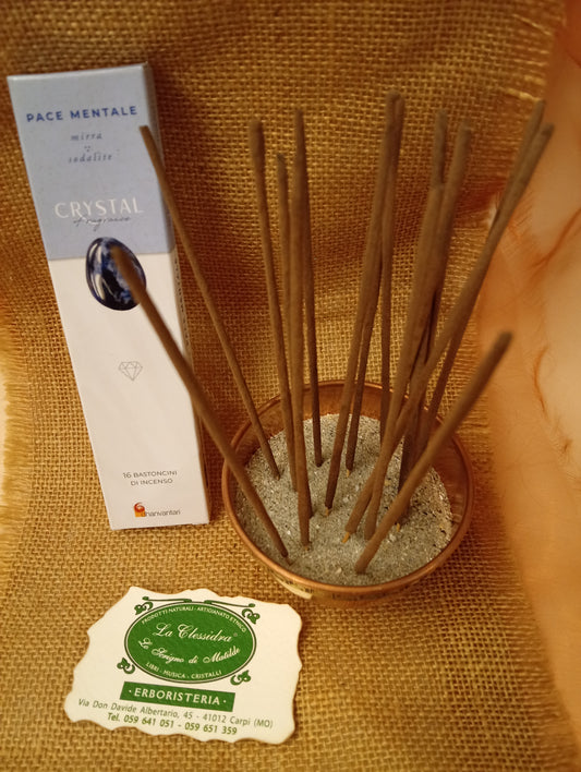 Confezione di Incenso Crystal Fragrance Pace Mentale Dhanvantari appoggiata accanto a una ciotola di rame con sedici bastoncini inseriti nella sabbia e al biglietto da visita dell’Erboristeria La Clessidra Carpi su sfondo in telo di yuta