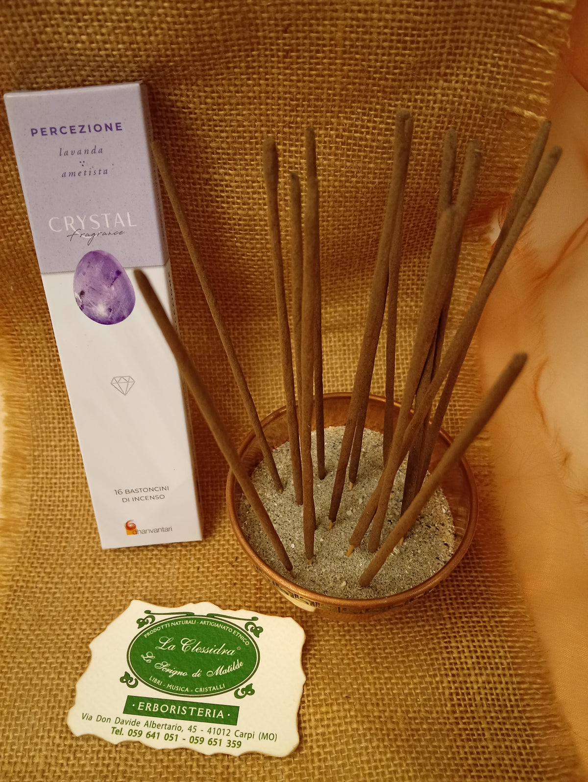 Confezione di Incenso Crystal Fragrance Percezione Dhanvantari appoggiata accanto a una ciotola di rame con sedici bastoncini inseriti nella sabbia e al biglietto da visita dell’Erboristeria La Clessidra Carpi su sfondo in telo di yuta.