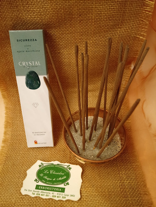 Confezione di Incenso Crystal Fragrance Sicurezza Dhanvantari appoggiata accanto a una ciotola di rame con sedici bastoncini inseriti nella sabbia e al biglietto da visita dell’Erboristeria La Clessidra Carpi su sfondo in telo di yuta