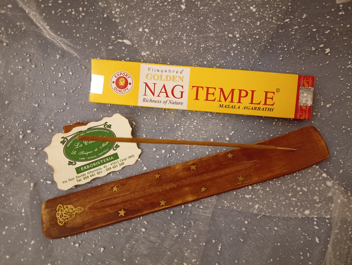 Confezione orizzontale di Incenso Golden Nag Temple Vijayshree appoggiata su barchetta di legno portaincenso con biglietto da visita dell’Erboristeria La Clessidra Carpi e sfondo con velo trasparente grigio azzurro puntinato di bianco