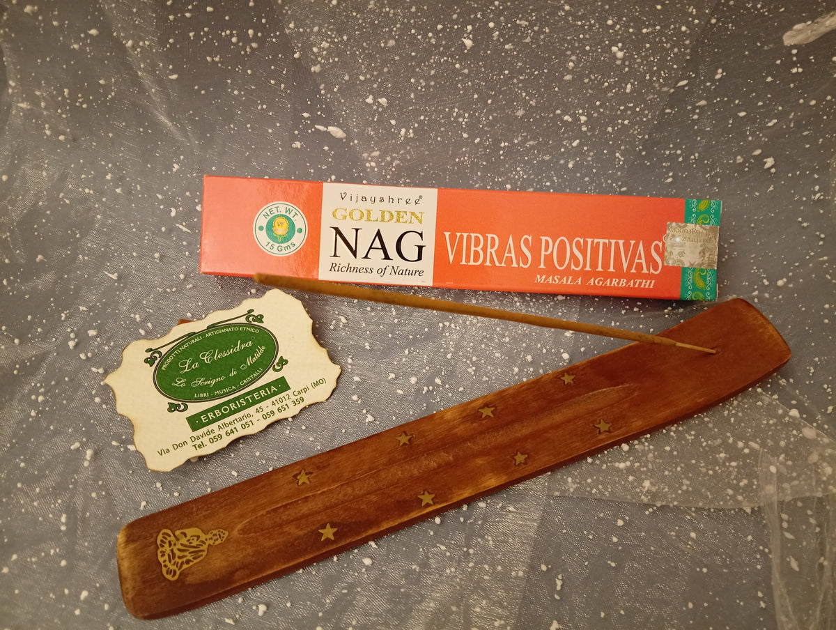 Confezione orizzontale di Incenso Golden Nag Vibras Positivas Vijayshree appoggiata su barchetta di legno portaincenso con biglietto da visita dell’Erboristeria La Clessidra Carpi e sfondo con velo trasparente grigio azzurro puntinato di bianco.
