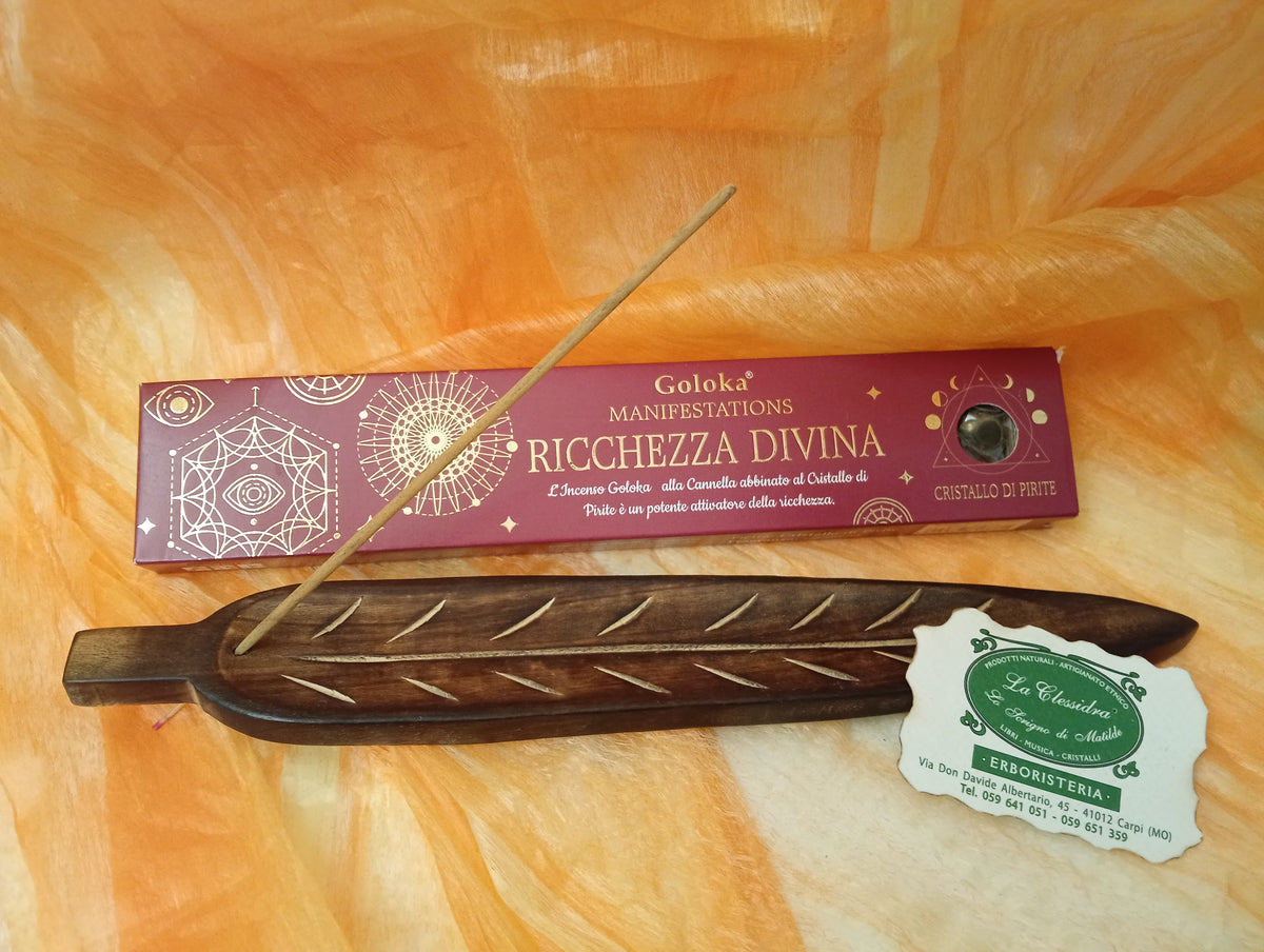 Confezione orizzontale di Incenso Goloka Manifestation Ricchezza Divina con Pirite burattata, davanti a un portaincenso artigianale a forma di foglia con stick, biglietto da visita dell’Erboristeria La Clessidra Carpi e sfondo con telo sfumato in toni aranciati orientali.