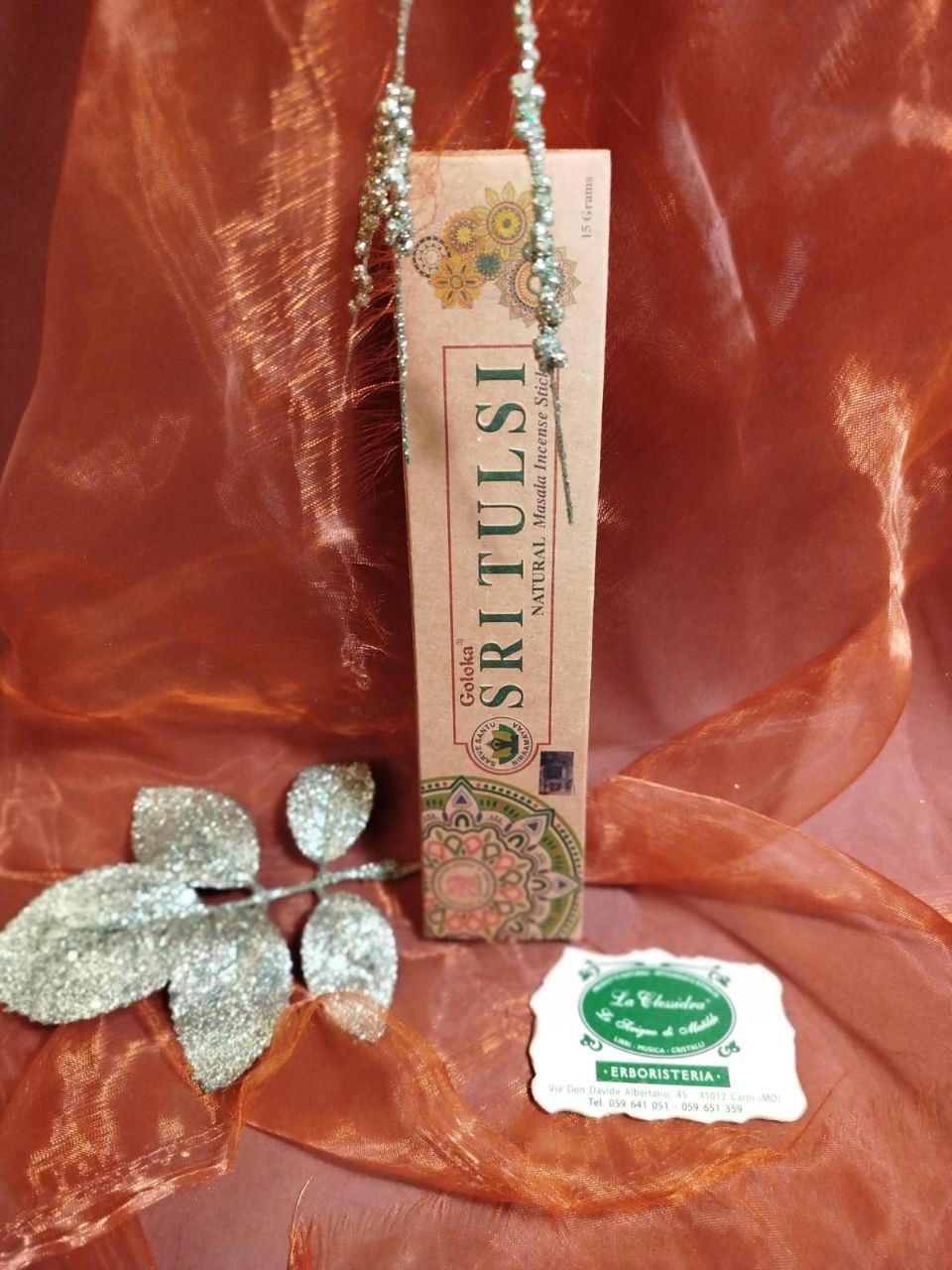 Incenso Goloka Natural Sri Tulsi su tessuto arancione con decorazioni argentate e biglietto del negozio La Clessidra a Carpi.