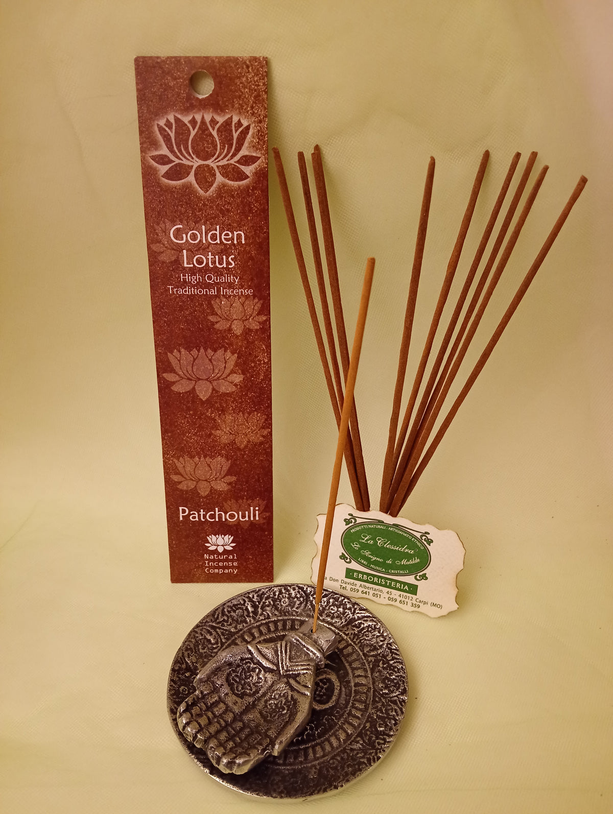 Incenso Patchouli Fiore d'Oriente Linea Golden Lotus, astuccio con mazzetto di bastoncini aperti in piedi, portaincenso in metallo con mano e biglietto da visita esposti da Erboristeria La Clessidra Carpi.