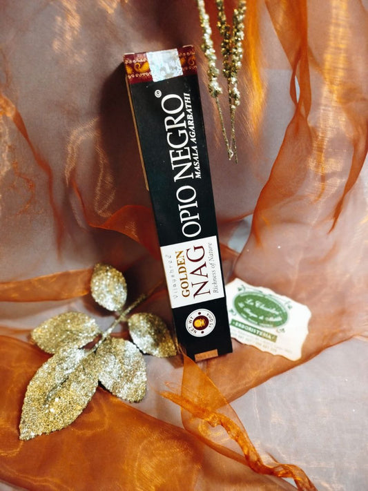 Confezione verticale di Incenso Golden Nag Opio Negro (Oppio Nero) Vijayshree circondata da foglie decorative con il biglietto da visita dell’Erboristeria La Clessidra Carpi e sfondo impreziosito da un velo trasparente color caldo orientale