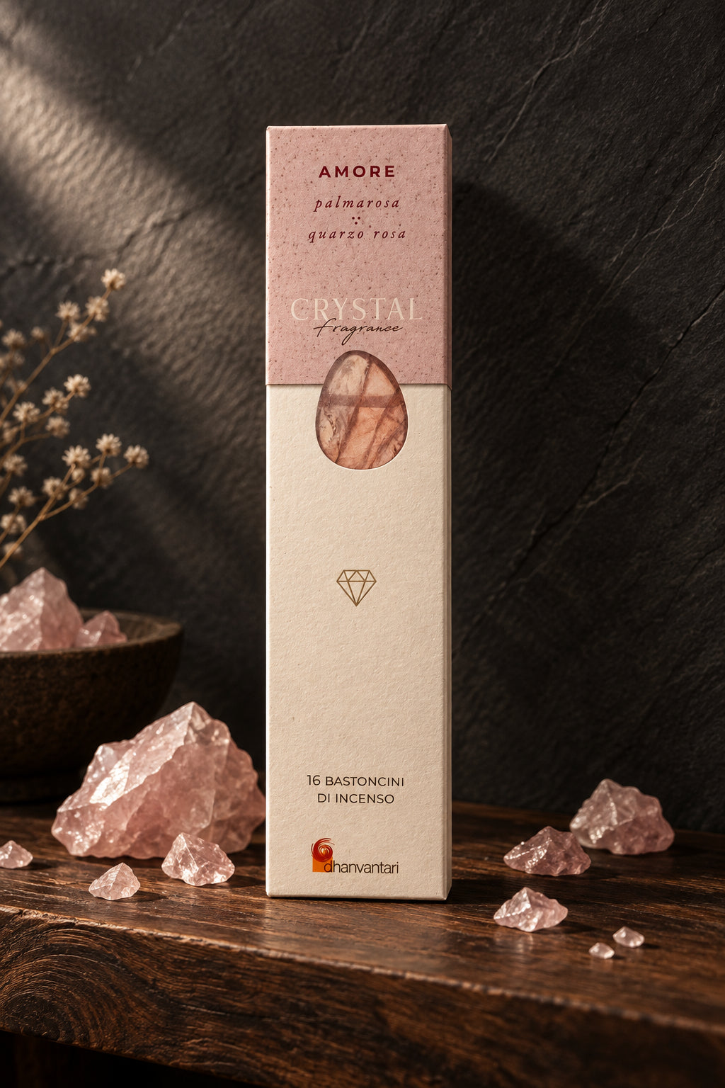 Astuccio rettangolare verticale di Incenso Crystal Fragrance Amore Dhanvantari su mensola di legno scuro davanti a una parete di ardesia con frammenti grezzi di pietra quarzo rosa, da Erboristeria La Clessidra Carpi.