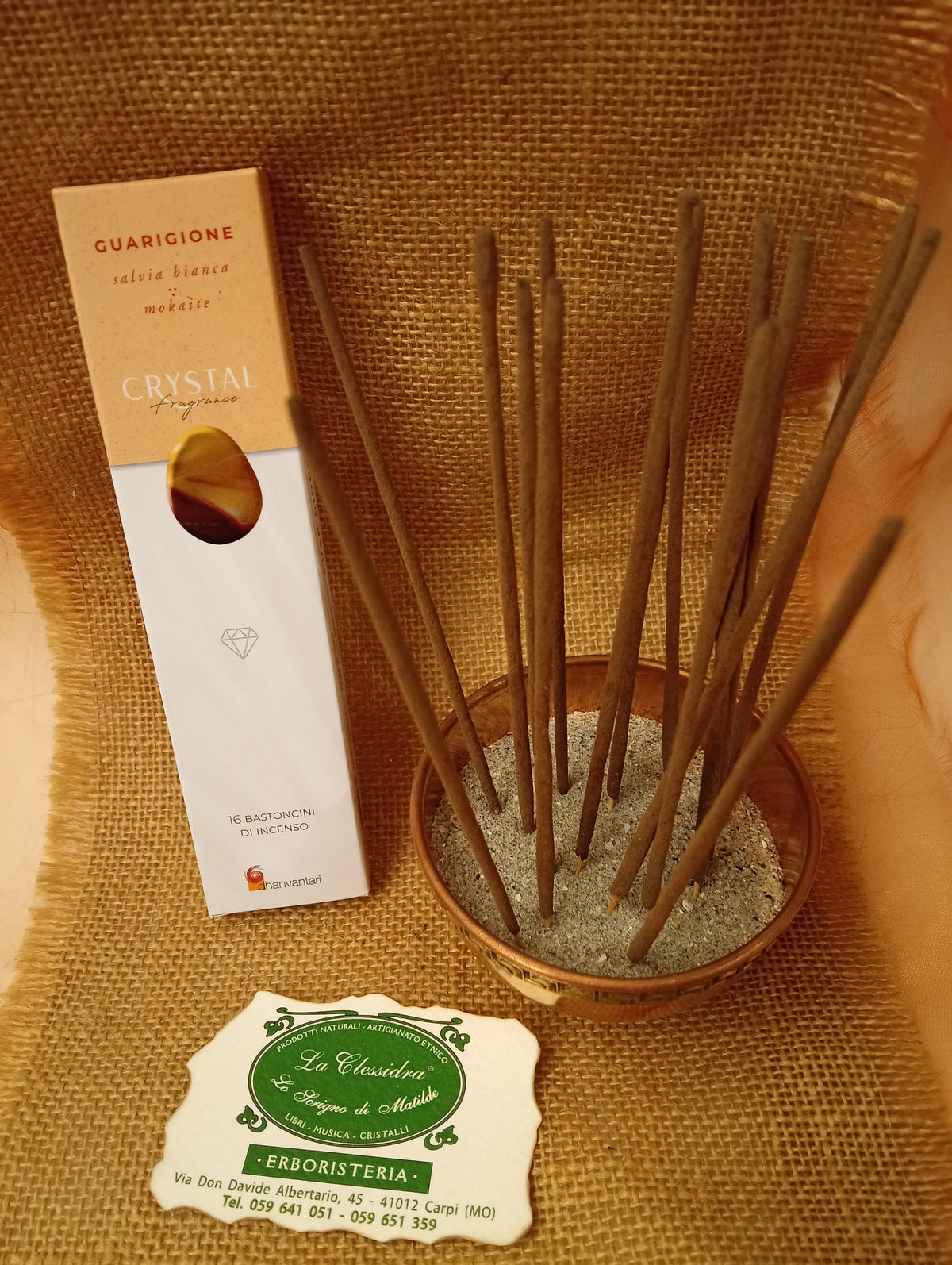 Confezione di Incenso Crystal Fragrance Guarigione Dhanvantari appoggiata accanto a una ciotola di rame con sedici bastoncini inseriti nella sabbia e al biglietto da visita dell’Erboristeria La Clessidra Carpi su sfondo in telo di yuta