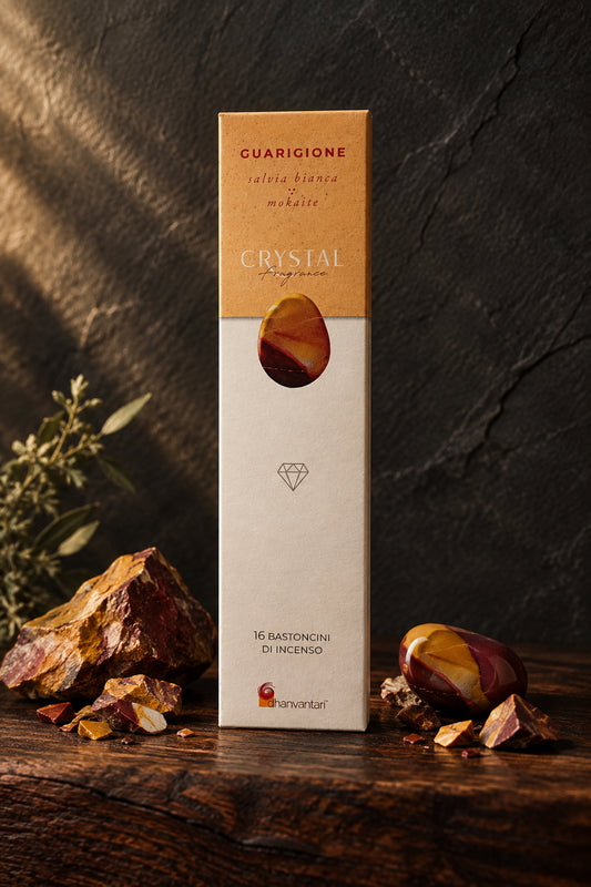 Astuccio rettangolare verticale di Incenso Crystal Fragrance Guarigione Dhanvantari su mensola di legno scuro davanti a una parete di ardesia con frammenti grezzi di pietra mokaite, da Erboristeria La Clessidra Carpi.