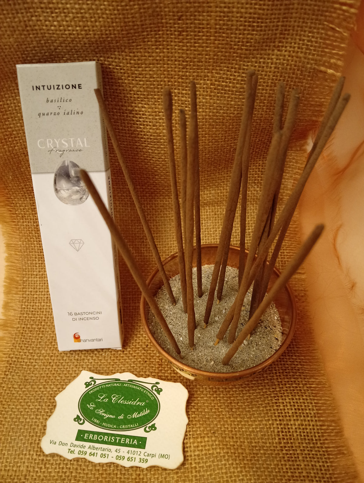 Confezione di Incenso Crystal Fragrance Intuizione Dhanvantari appoggiata accanto a una ciotola di rame con sedici bastoncini inseriti nella sabbia e al biglietto da visita dell’Erboristeria La Clessidra Carpi su sfondo in telo di yuta