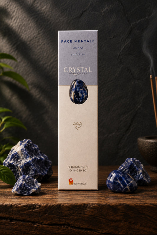 Astuccio rettangolare verticale di Incenso Crystal Fragrance Pace Mentale Dhanvantari su mensola di legno scuro davanti a una parete di ardesia con frammenti grezzi di pietra sodalite blu, da Erboristeria La Clessidra Carpi.