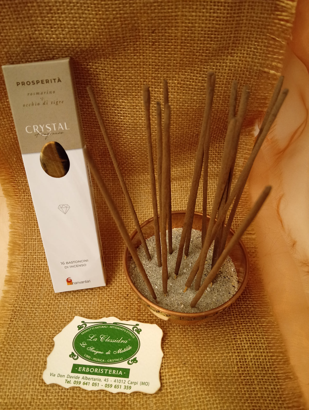Confezione di Incenso Crystal Fragrance Prosperità Dhanvantari appoggiata accanto a una ciotola di rame con sedici bastoncini inseriti nella sabbia e al biglietto da visita dell’Erboristeria La Clessidra Carpi su sfondo in telo di yuta