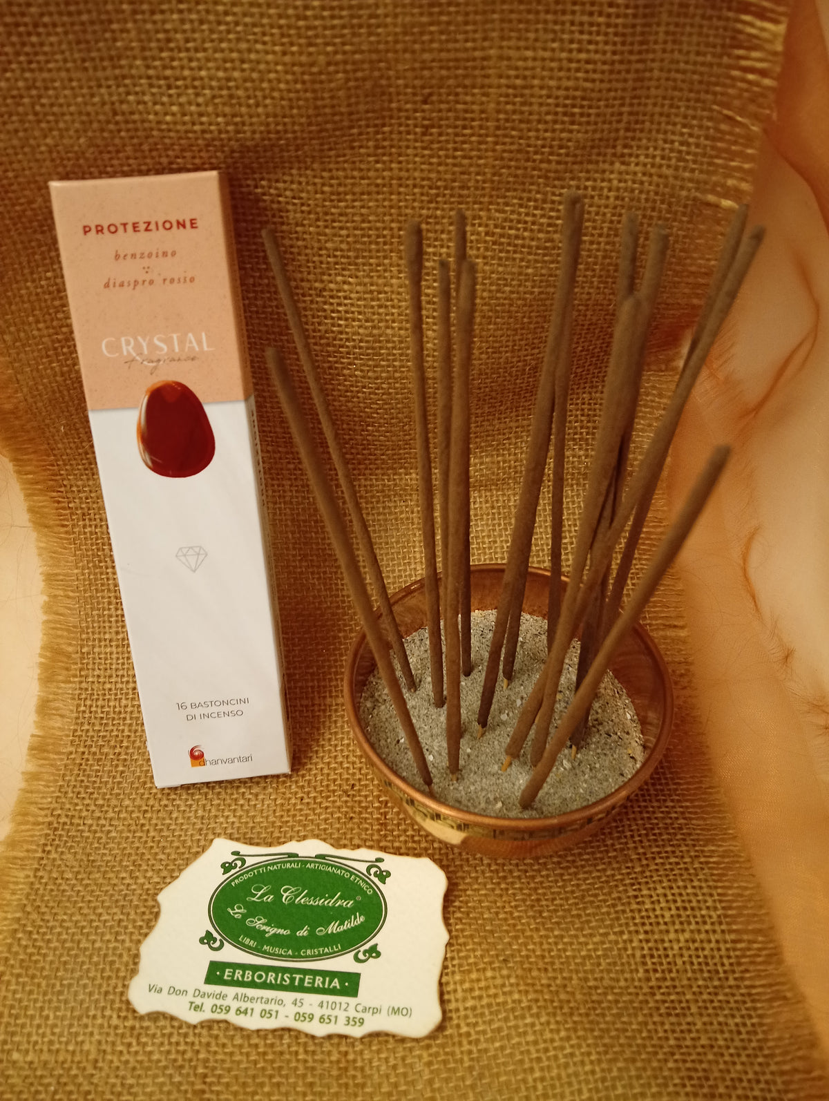 Confezione di Incenso Crystal Fragrance Protezione Dhanvantari appoggiata accanto a una ciotola di rame con sedici bastoncini inseriti nella sabbia e al biglietto da visita dell’Erboristeria La Clessidra Carpi su sfondo in telo di yuta