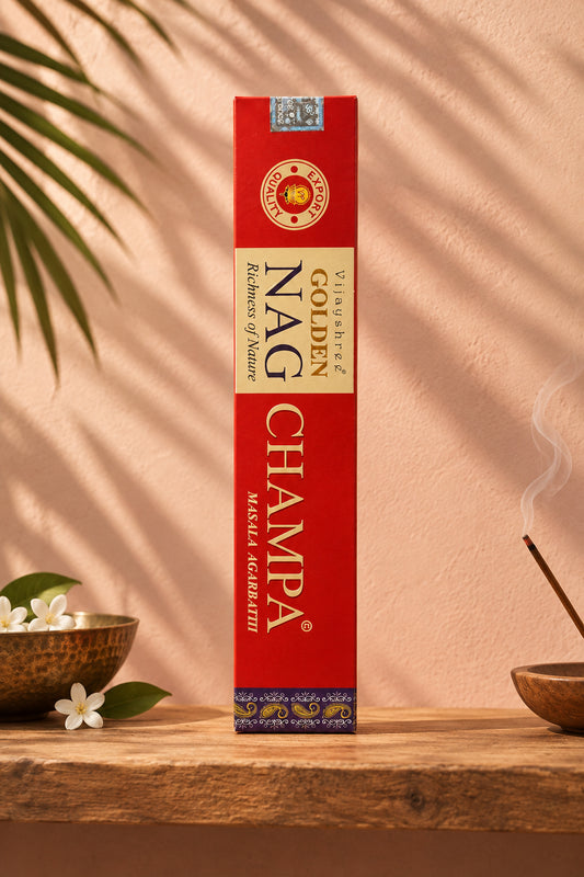 Astuccio rettangolare di Incenso Golden Nag Champa Vijayshree rosso e oro in verticale su mensola di legno grezzo davanti a una parete di intonaco color mattone rosato con l'ombra sfumata di una foglia di palma, da Erboristeria La Clessidra Carpi
