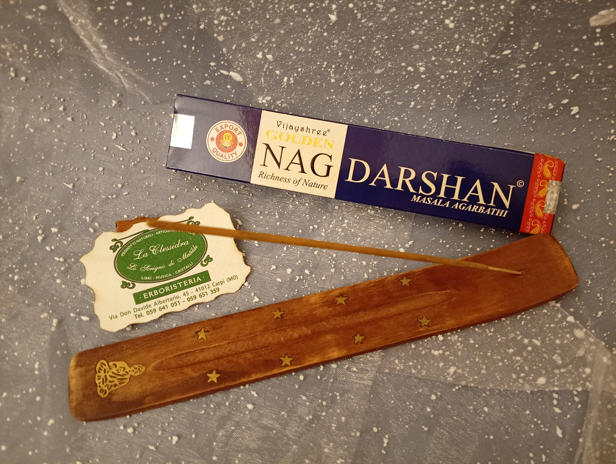 Confezione orizzontale di Incenso Golden Nag Darshan Vijayshree appoggiata su barchetta di legno portaincenso con biglietto da visita dell’Erboristeria La Clessidra Carpi e sfondo con velo trasparente grigio azzurro puntinato di bianco.