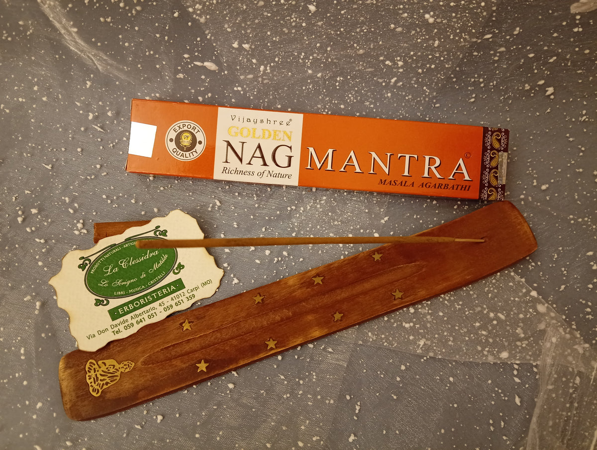 Confezione orizzontale di Incenso Golden Nag Mantra Vijayshree appoggiata su barchetta di legno portaincenso con biglietto da visita dell’Erboristeria La Clessidra Carpi e sfondo con velo trasparente grigio azzurro puntinato di bianco