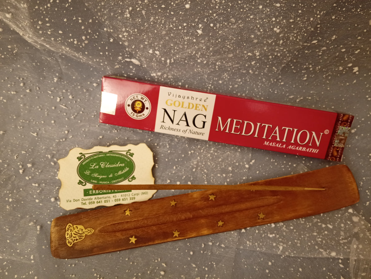 Confezione orizzontale di Incenso Golden Nag Meditation Vijayshree appoggiata su barchetta di legno portaincenso con biglietto da visita dell’Erboristeria La Clessidra Carpi e sfondo con velo trasparente grigio azzurro puntinato di bianco