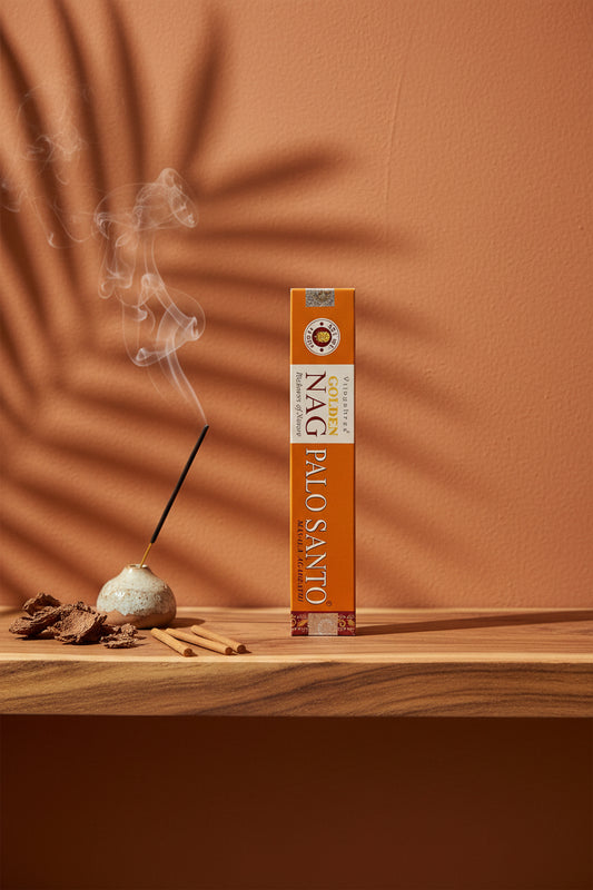 Astuccio di Incenso Golden Nag Palo Santo Vijayshree su mensola di legno grezzo davanti a parete terracotta con ombre di foglie di palma, portaincenso artigianale con stick e scaglie di corteccia, da Erboristeria La Clessidra Carpi