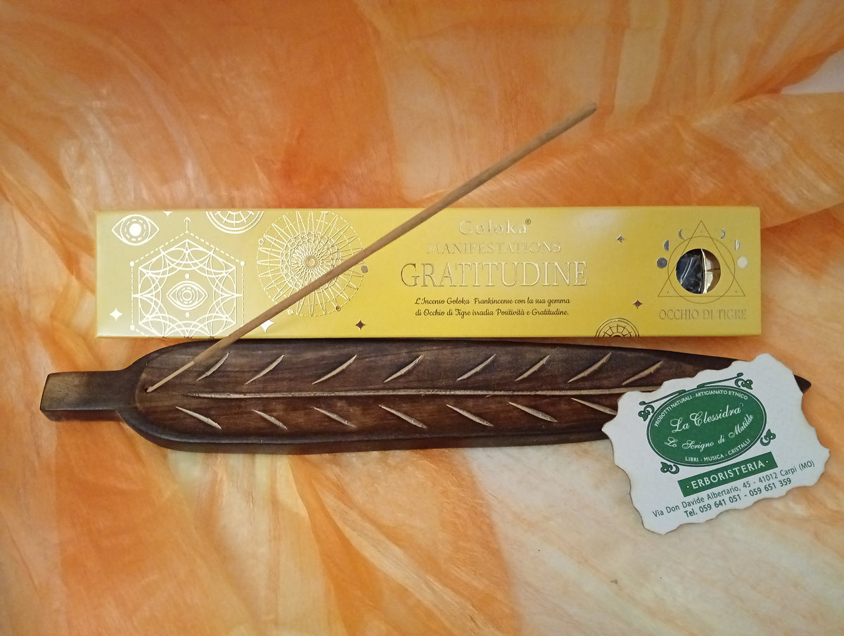 Confezione orizzontale di Incenso Goloka Manifestation Gratitudine con Occhio di Tigre burattato, davanti a un portaincenso artigianale a forma di foglia con stick, biglietto da visita dell’Erboristeria La Clessidra Carpi e sfondo con telo sfumato in toni aranciati orientali