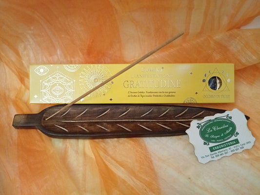 Confezione orizzontale di Incenso Goloka Manifestation Gratitudine con Occhio di Tigre burattato, davanti a un portaincenso artigianale a forma di foglia con stick, biglietto da visita dell’Erboristeria La Clessidra Carpi e sfondo con telo sfumato in toni aranciati orientali