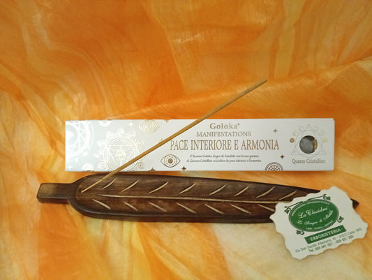 Confezione orizzontale di Incenso Goloka Manifestation Pace Interiore e Armonia con Quarzo Cristallino, davanti a un portaincenso artigianale a forma di foglia con stick, biglietto da visita dell’Erboristeria La Clessidra Carpi e sfondo con telo sfumato in toni aranciati orientali.