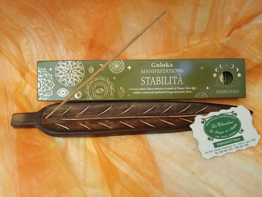 Confezione orizzontale di Incenso Goloka Manifestation Stabilità con Diaspro Rosso burattato, davanti a un portaincenso artigianale a forma di foglia con stick, biglietto da visita dell’Erboristeria La Clessidra Carpi e sfondo con telo sfumato in toni aranciati orientali.