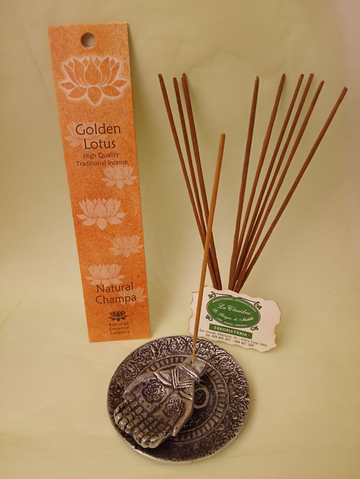 Incenso Natural Champa Fiore d'Oriente Linea Golden Lotus, astuccio con mazzetto di bastoncini aperti in piedi, portaincenso in metallo con mano e biglietto da visita esposti da Erboristeria La Clessidra Carpi.