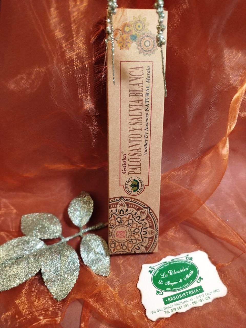 Incenso Goloka Natural Palo Santo e Salvia Bianca su tessuto arancione con decorazioni argentate e biglietto del negozio La Clessidra a Carpi.
