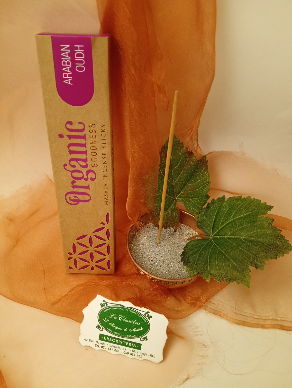Astuccio in verticale di Incenso Organic Goodness Arabian Oudh Song of India accanto a ciotola rotonda in ottone con sabbia, uno stick, due foglie di vite e biglietto da visita verde su sfondo con velo arancio marrone