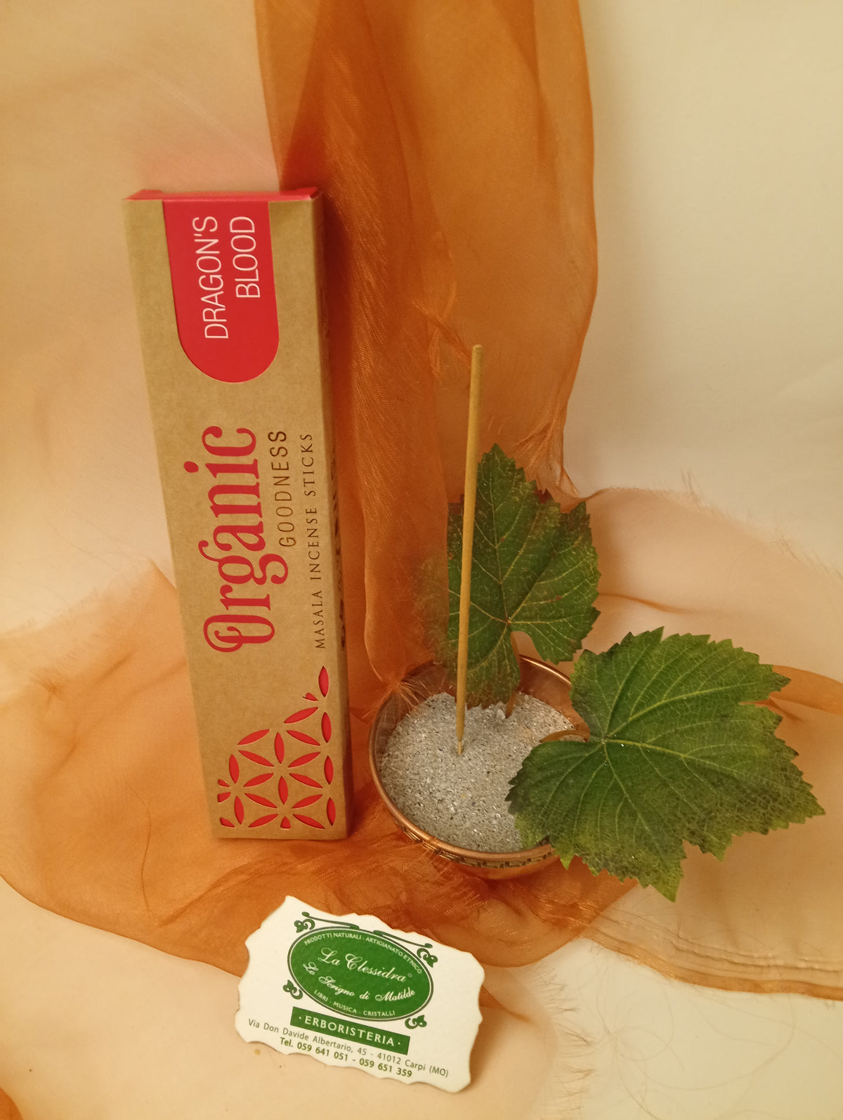 Astuccio in verticale di Incenso Organic Goodness Dragon's Blood Song of India accanto a ciotola rotonda in ottone con sabbia, uno stick, due foglie di vite e biglietto da visita verde su sfondo con velo arancio marrone