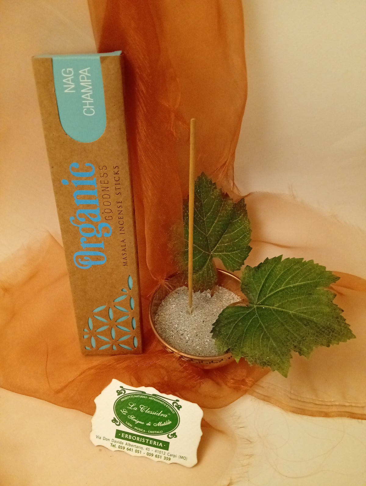 Astuccio in verticale di Incenso Organic Goodness Nag Champa Song of India accanto a ciotola rotonda in ottone con sabbia, uno stick, due foglie di vite e biglietto da visita verde su sfondo con velo arancio marrone