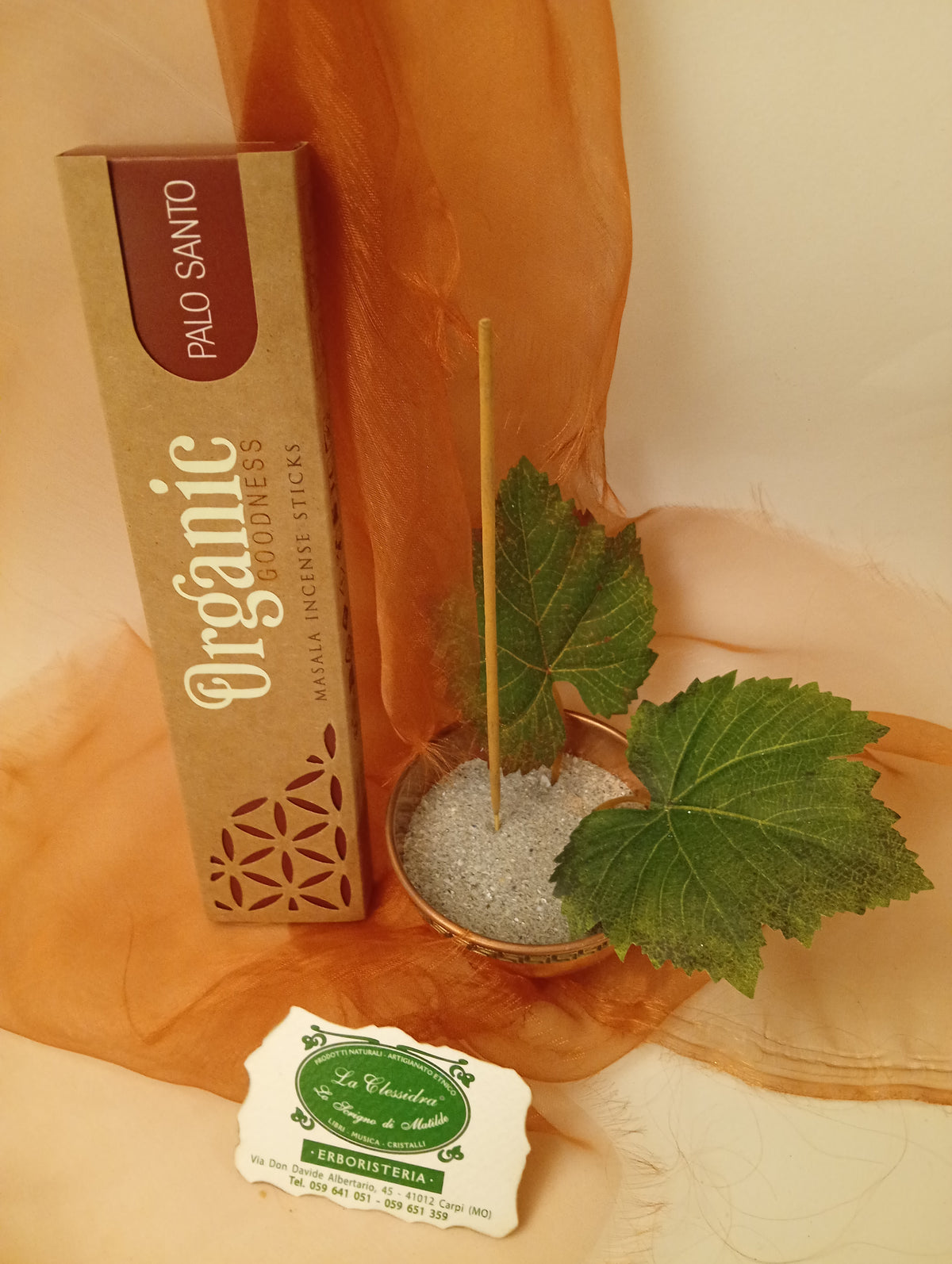 Astuccio in verticale di Incenso Organic Goodness Palo Santo Song of India accanto a ciotola rotonda in ottone con sabbia, uno stick, due foglie di vite e biglietto da visita verde su sfondo con velo arancio marrone