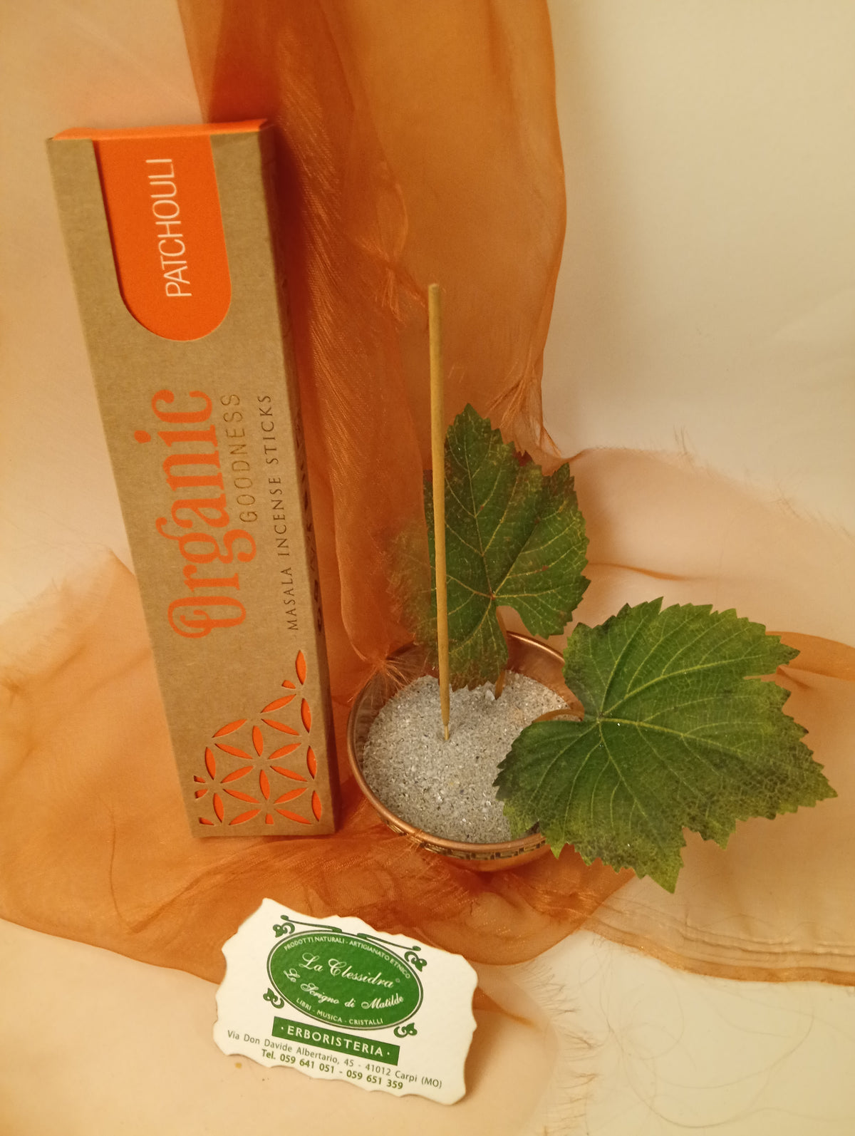 Astuccio in verticale di Incenso Organic Goodness Patchouli Song of India accanto a ciotola rotonda in ottone con sabbia, uno stick, due foglie di vite e biglietto da visita verde su sfondo con velo arancio marrone