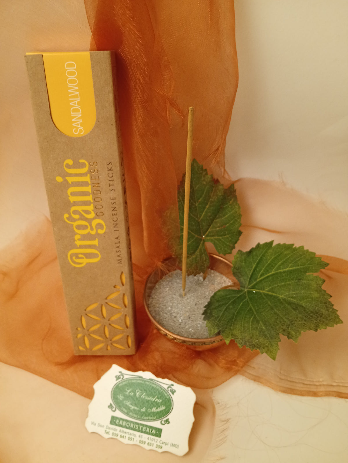 Astuccio in verticale di Incenso Organic Goodness Sandalwood Song of India accanto a ciotola rotonda in ottone con sabbia, uno stick, due foglie di vite e biglietto da visita verde su sfondo con velo arancio marrone