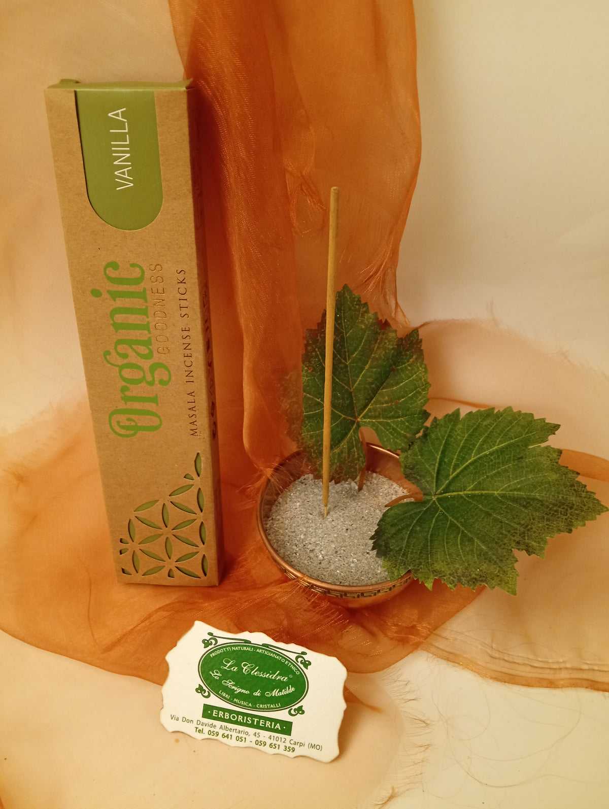 Astuccio in verticale di Incenso Organic Goodness Vanilla Song of India accanto a ciotola rotonda in ottone con sabbia, uno stick, due foglie di vite e biglietto da visita verde su sfondo con velo arancio marrone