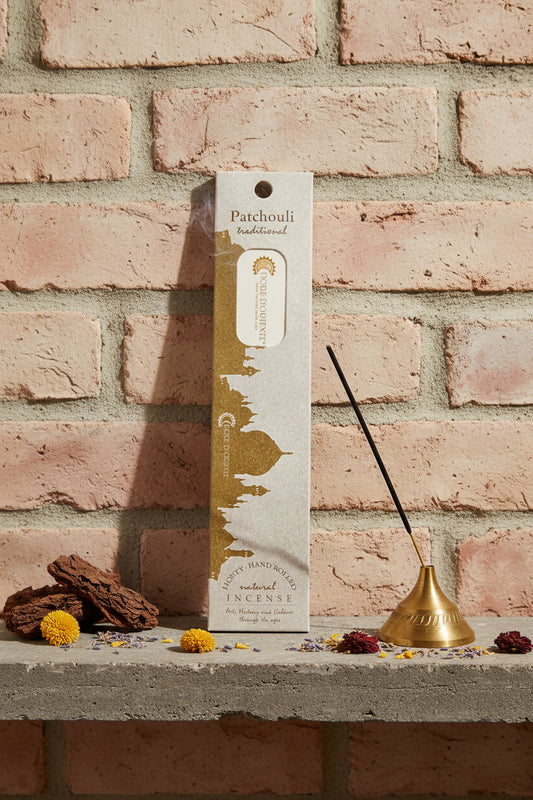 Incenso Patchouli Fiore d'Oriente Linea Traditional con stick acceso in portaincenso di ottone, cortecce e fiori su base di pietra e muro antico, da Erboristeria La Clessidra Carpi.