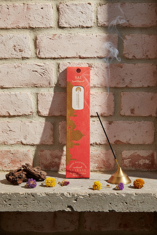 Incenso Sai Fiore d'Oriente Linea Traditional con stick acceso in portaincenso di ottone, cortecce e fiori su base di pietra e muro antico, da Erboristeria La Clessidra Carpi.
