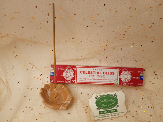 Confezione orizzontale di Incenso Celestial Bliss Satya con un portaincenso in terracotta a forma di due mani unite con il biglietto da visita dell’Erboristeria La Clessidra Carpi davanti a uno sfondo con velo trasparente a punti color oro