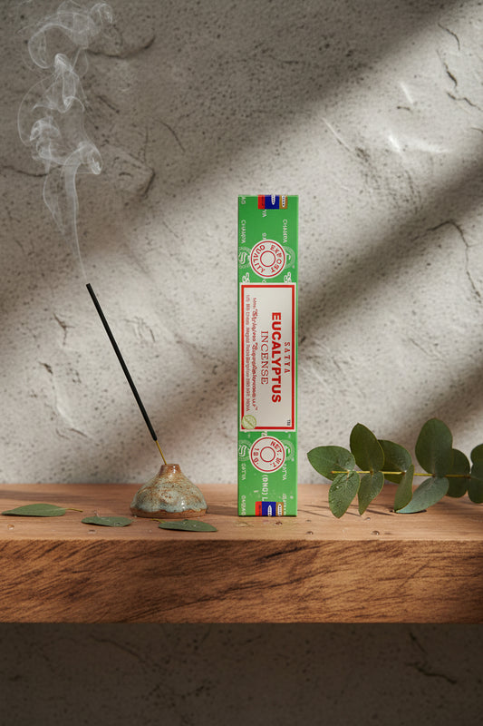 Astuccio di Incenso Eucalyptus (Eucalipto) Satya su mensola di legno grezzo davanti a parete di roccia grigio chiara con luce laterale, portaincenso artigianale con stick e foglie fresche di eucalipto bagnate di rugiada, da Erboristeria La Clessidra Carpi.