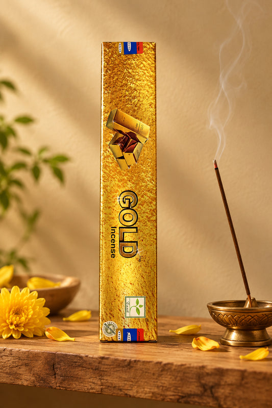 Astuccio rettangolare verticale di Incenso Gold Satya color oro su mensola di legno grezzo davanti a una parete di intonaco beige sabbia con una luce soffusa e sfumata sul retro e petali di fiori dorati sparsi, da Erboristeria La Clessidra Carpi.