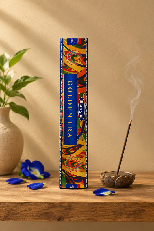 Astuccio rettangolare verticale di Incenso Golden Era Satya con dettagli blu elettrico su mensola di legno grezzo davanti a una parete di intonaco beige sabbia con una luce soffusa e sfumata sul retro e petali di fiori blu sparsi, da Erboristeria La Clessidra Carpi
