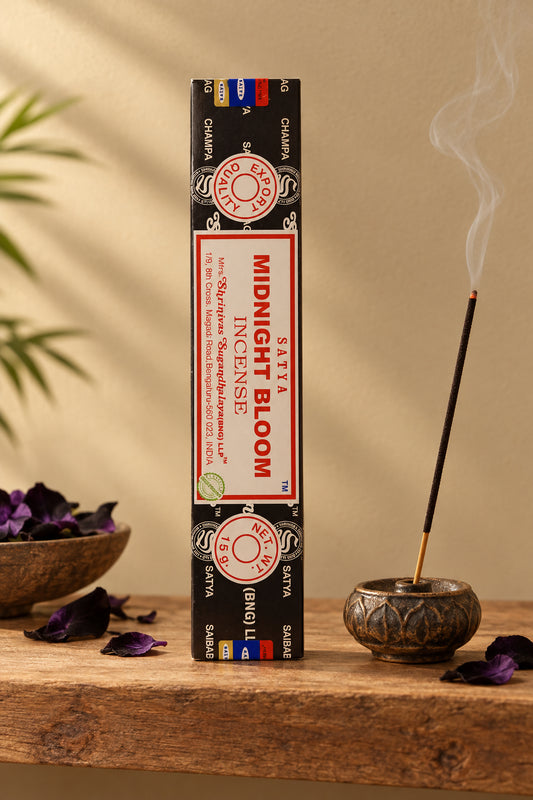 Astuccio rettangolare verticale di Incenso Midnight Bloom Satya nero e oro su mensola di legno grezzo davanti a una parete di intonaco beige sabbia con una luce soffusa e sfumata sul retro e petali di fiori neri e viola sparsi, da Erboristeria La Clessidra Carpi.