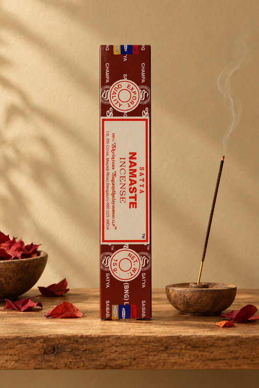 Astuccio rettangolare verticale di Incenso Namastè Satya rosso scuro marroncino su mensola di legno grezzo davanti a una parete di intonaco beige sabbia con una luce soffusa e sfumata sul retro e petali di fiori color rosso mattone sparsi, da Erboristeria La Clessidra Carpi