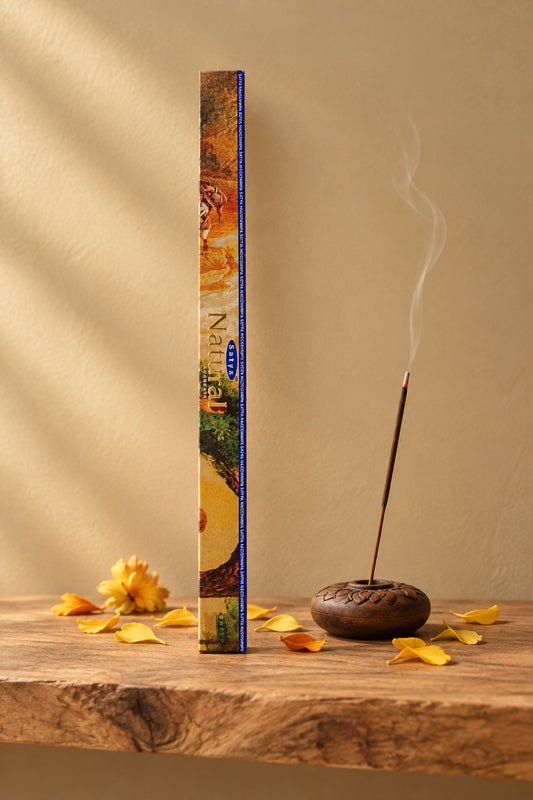 Confezione quadrata di Incenso Natural Satya astuccio quadro giallo oro e ocra su mensola di legno grezzo davanti a una parete di intonaco beige sabbia con una luce soffusa e sfumata sul retro e petali dorati sparsi, da Erboristeria La Clessidra Carpi