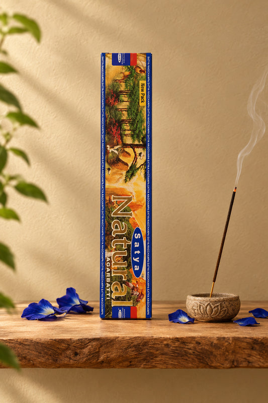 Astuccio rettangolare verticale di Incenso Natural Satya con scritte blu su mensola di legno grezzo davanti a una parete di intonaco beige sabbia con una luce soffusa e sfumata sul retro e petali di fiori blu sparsi, da Erboristeria La Clessidra Carpi.