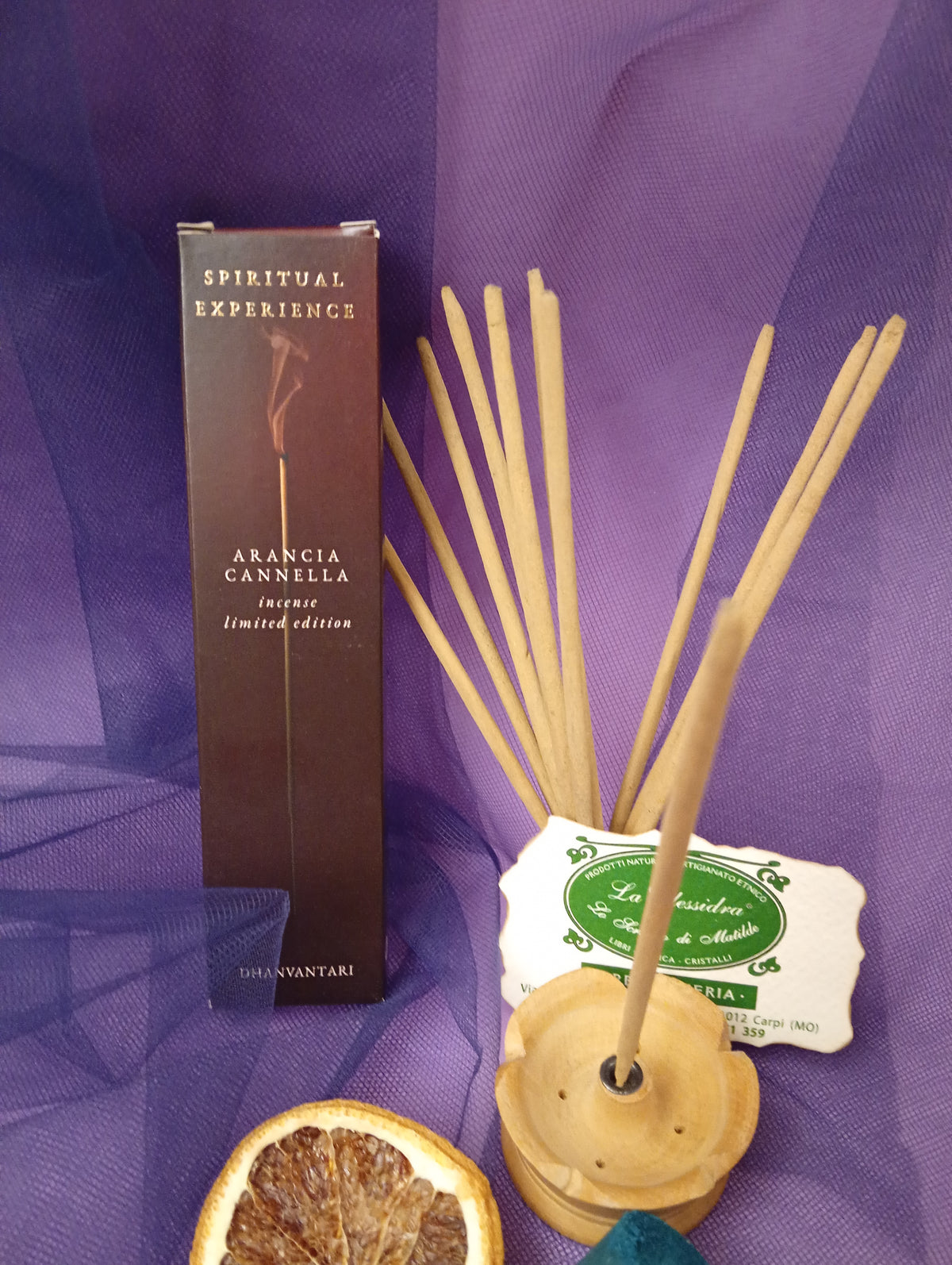 Astuccio di Incenso Spiritual Experience Arancia Cannella Dhanvantari in verticale con mazzo di bastoncini inseriti nel portaincenso in legno chiaro, fetta di arancia essiccata, legnetti e pietra azzurra su sfondo con velo viola, Erboristeria La Clessidra Carpi