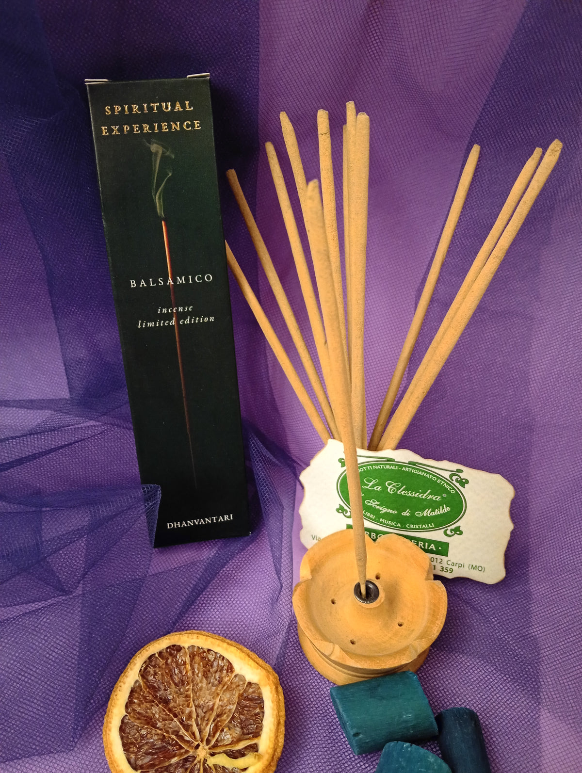 Astuccio di Incenso Spiritual Experience Balsamico Dhanvantari in verticale con mazzo di bastoncini inseriti nel portaincenso in legno chiaro, fetta di arancia essiccata, legnetti e pietra azzurra su sfondo con velo viola, Erboristeria La Clessidra Carpi