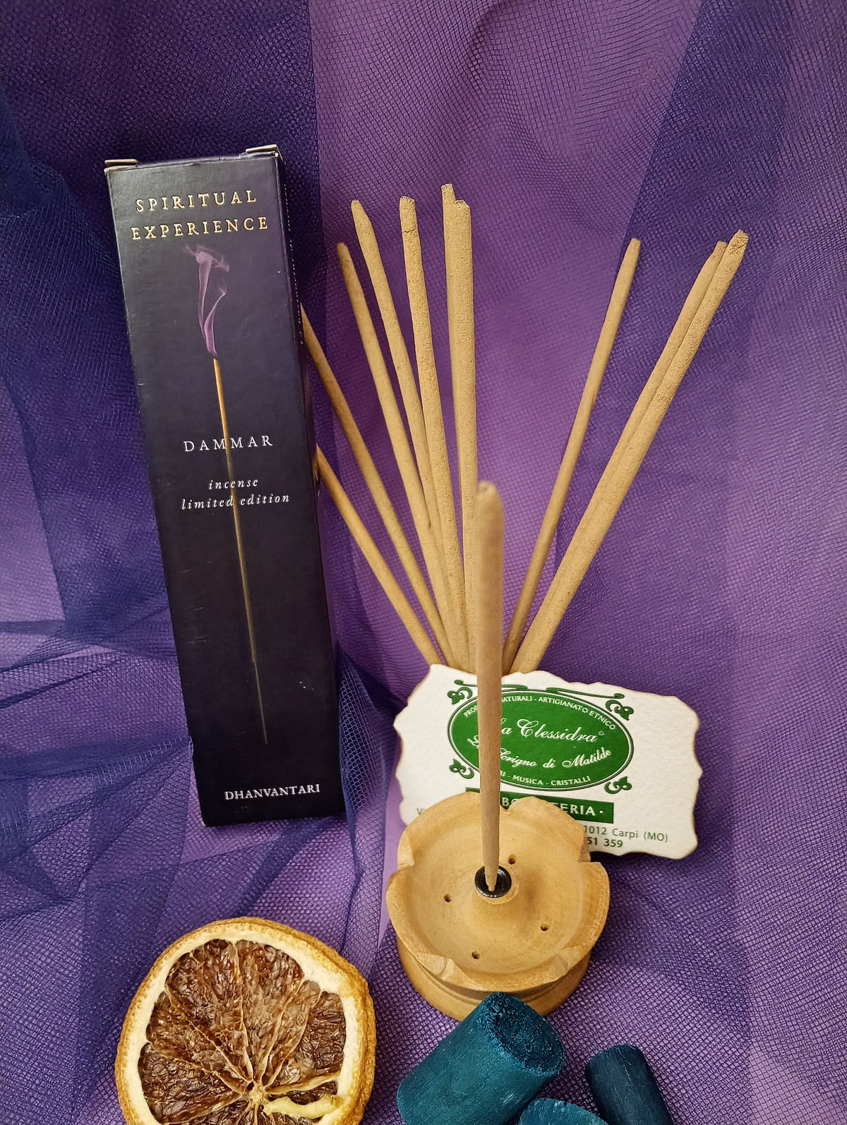 Astuccio di Incenso Spiritual Experience Dammar Dhanvantari in verticale con mazzo di bastoncini inseriti nel portaincenso in legno chiaro, fetta di arancia essiccata, legnetti e pietra azzurra su sfondo con velo viola, Erboristeria La Clessidra Carpi